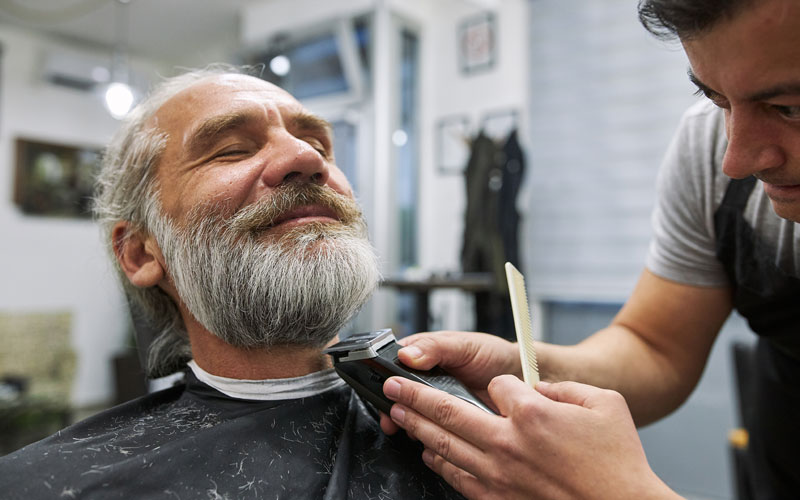 A barber trims an older mans beard in a salon with a comb and electric trimmer.