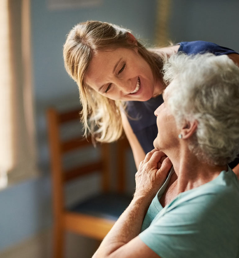 Caregiver smiling and interacting with a seated elderly woman in a warmly lit room.