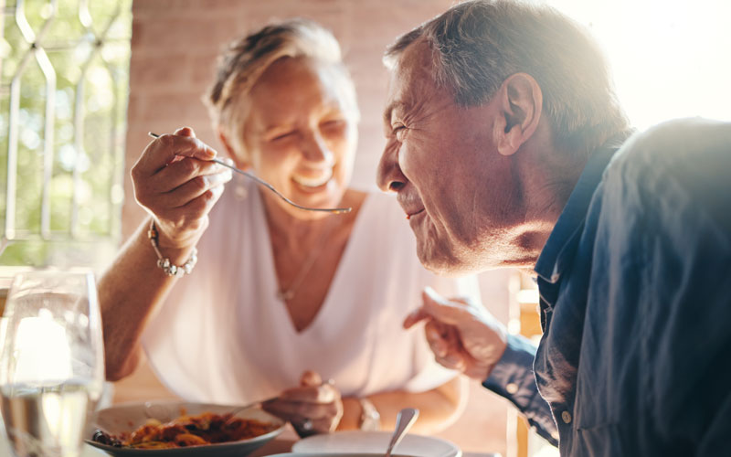 Elderly woman feeding elderly man with a spoon in a sunlit dining setting, both smiling warmly.