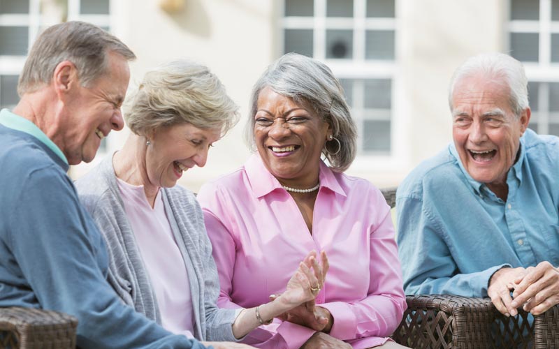 Elderly friends laughing and enjoying outdoor conversation in a sunny courtyard.