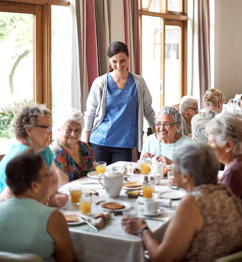 Elderly residents dining together while a caregiver in blue scrubs smiles beside them.