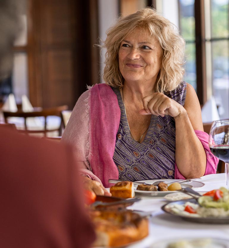 Elderly woman enjoying a meal with her friends at a restaurant, smiling and wearing a purple patterned top