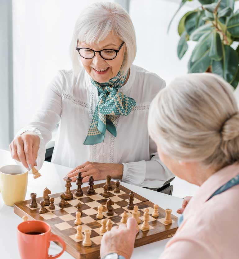 Two elderly women smiling and playing chess at a table with coffee mugs in a bright room.