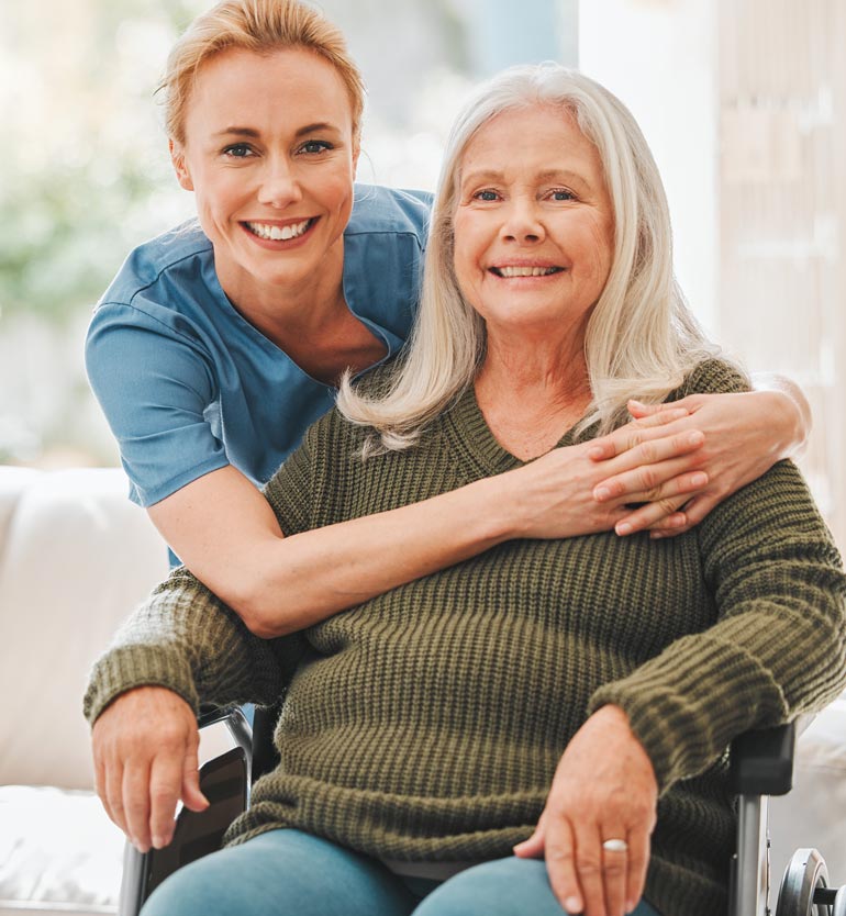 Smiling healthcare worker hugging a happy senior woman in a wheelchair in a well-lit room.