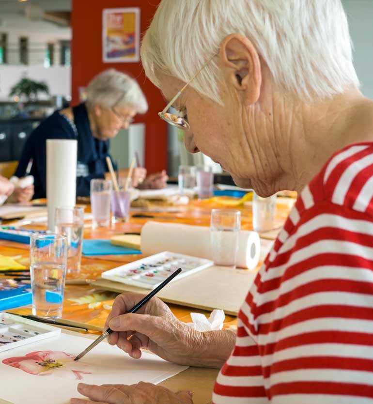 Elderly woman painting with watercolor in an art class with other seniors in the background