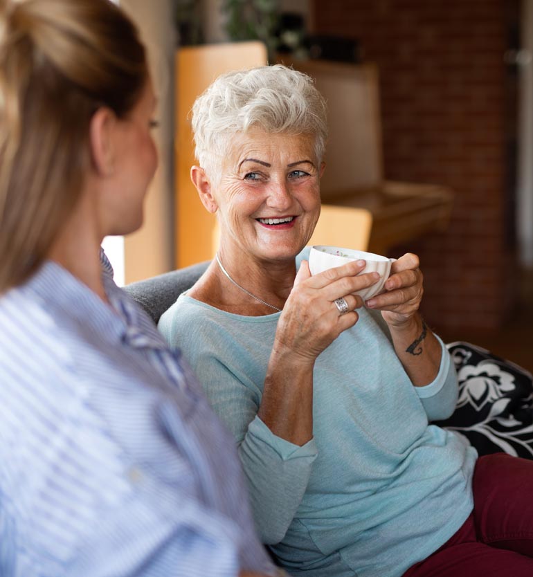 Senior woman smiling and holding a cup while talking with a younger woman on a sofa.