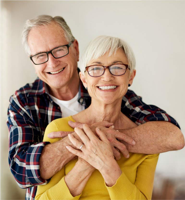 Senior couple embracing and smiling while standing together, both wearing glasses and casual clothing.
