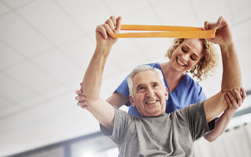 A senior man smiling while exercising with a stretch band, assisted by a caregiver in a bright room.