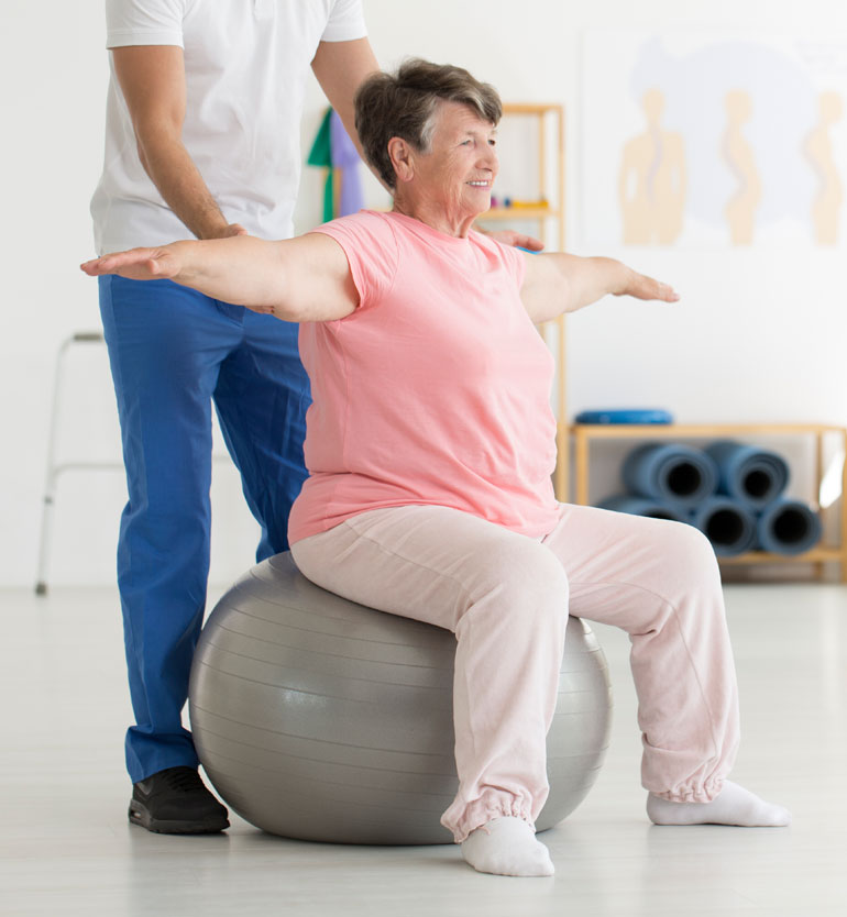 A senior woman sits on an exercise ball, guided by a therapist during a physical therapy session