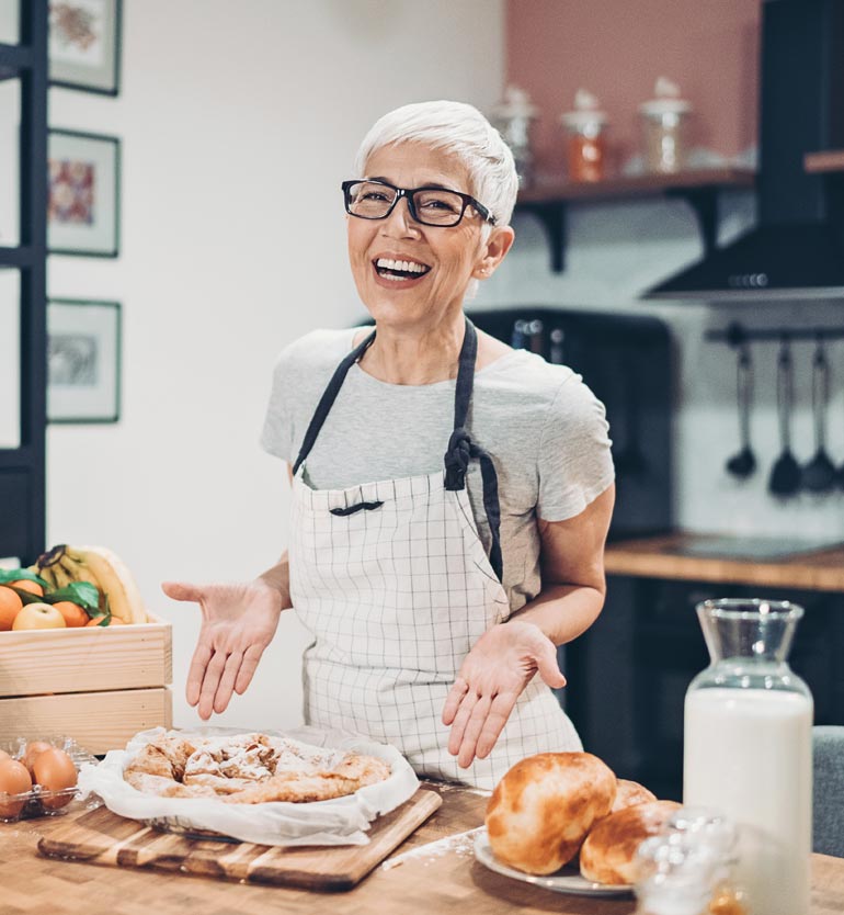 Senior woman with glasses and apron baking in a kitchen, showing off a freshly baked dish.