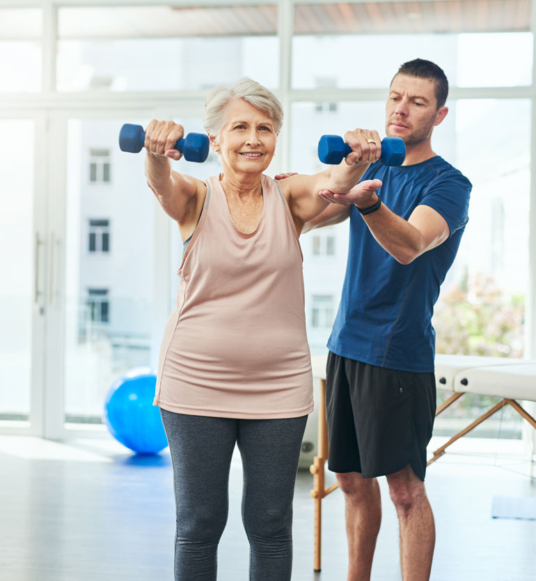 Senior woman lifting weights with a trainer assisting in a bright, modern gym setting.