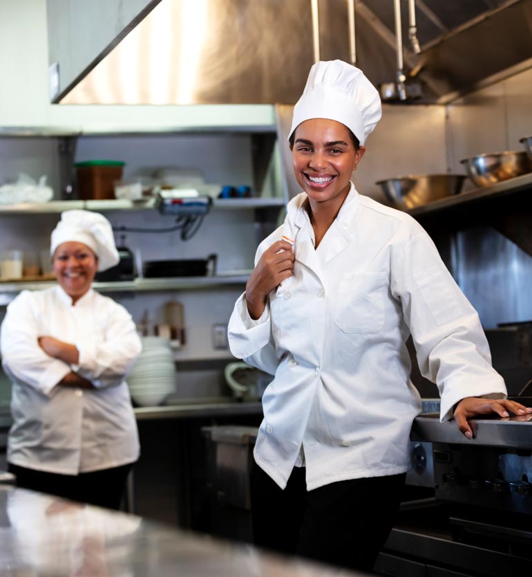 Two chefs in a commercial kitchen, one in the foreground and one in the background, both smiling.